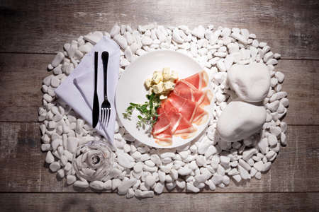 A white plate with hamon, blue cheese on white stones and on a wooden background. A transparent, empty glass near the food composition. Romantic dinner at the restaurant.の写真素材