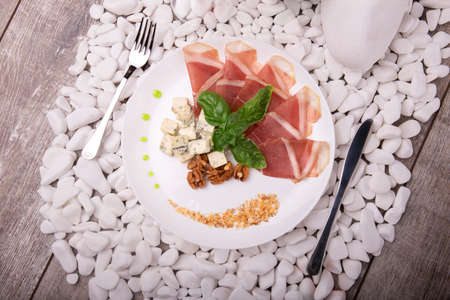 A white plate with prosciutto, blue cheese on white stones and on a wooden background. A composition of hamon, walnuts, basil and sauce on a plate. Romantic dinner at the restaurant.の写真素材