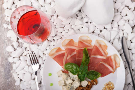 A plate with prosciutto, basil, and blue cheese on a white stone background. A glass of semi-sweet bright red wine near the expensive dish. Business dinner at the restaurant.の写真素材