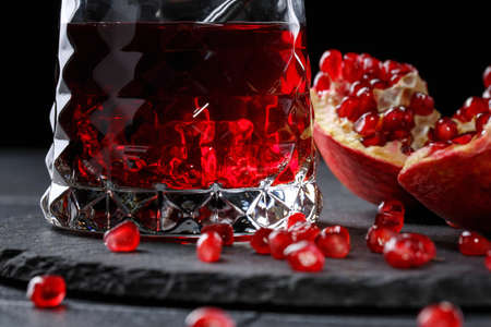Macro picture of a transparent glass full of sweet pomegranate juice with cubes of ice on a saturated black background. The ripe garnet broken into pieces on a table. Healthy and organic ingredients.の写真素材