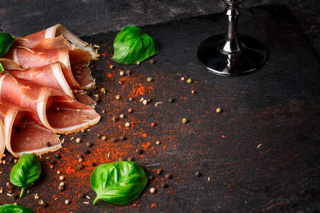 Close-up picture of sliced prosciutto and green basil leaves on a black stone background. Tasty snacks for alcoholic drinks.の写真素材
