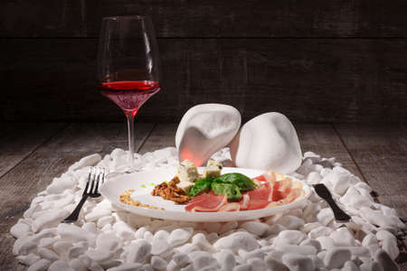 A luxurious composition of a glass of wine and restaurant lunch on a plate on a pile of white rocks. Walnuts, ham and white cheese with fork and knife on a brown wooden background.の写真素材