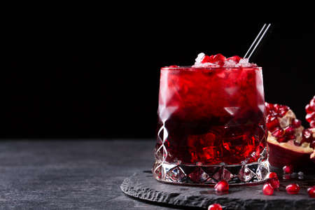 Close-up of a transparent glass of sweet pomegranate juice with ice and a cut garnet on a round plate on a saturated black background. Healthful and fruity red cocktail with straws and fresh garnet.の写真素材