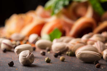 A close-up picture of light brown pistachios with black pepper, basil leaf and cut ham on a black blurred stone table background. Nutritious bar appetizers. Nuts full of vitamins.の写真素材