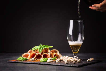 A man pours intoxicating beer into a transparent glass on a saturated black background. An Italian drying balyk in thin slices with green basil near the glass. Prosciutto and pistachios on the table.の写真素材