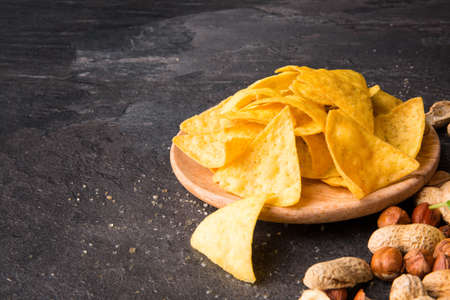 A view from above on gold nacho chips on a light wooden plate. Spicy snacks with salty peanuts and peeled hazelnuts. Mexican traditional appetizer on a black table background. Copy space.の写真素材