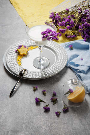 Organic yogurt in a transparent margarita glass on a plate on a colorful background. Bright yellow and blue fabric and purple summer flowers next to a natural, fresh and white milkshake.の写真素材