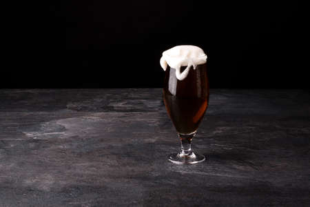 A big glass of beer full with brown ale and with white foam blowed away on a dark stone background.