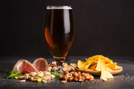 Closeup of moist glass of brown ale, with crisps, bacon, peanuts, hazelnuts, and pistachios on a dark background. Snacks for alcohol drink.の写真素材