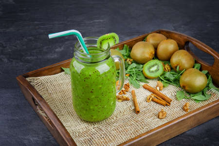 Close-up of a refreshing kiwi drink on a gray stone background. A cocktail in a mason jar with a colorful straw. Colorful kiwis, cinnamon sticks and mint leaves next to a cool drink on a wooden tray.の写真素材