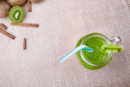 Top view of a mason jar full of kiwi smoozie with drinking straw and a slice of kiwi on a light wooden background. A heap of cinnamon and kiwis.の写真素材