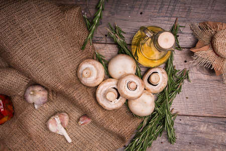 A view from above of a heap of fresh white mushrooms from a forest on a wooden background. A vase full of sunflowers oil, green rosemary and spicy garlic on an old brown bag on the table.の写真素材