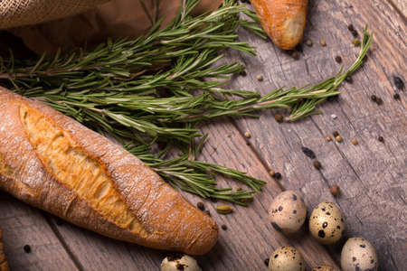 Closeup of wheat baguette, twigs of fragrant rosemary, speckled quail eggs, seasonings on a light wooden background.の写真素材
