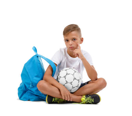 A boy sitting in the lotus position. A sportive kid with bright satchel and soccer ball isolated on a white background.の写真素材