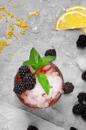 A view from above on a composition of glass of icy beverage with peppermint and blackberries on a gray background. Ripe, fresh blackberries, bright mint and a cut yellow lemon for a cold drink.の写真素材