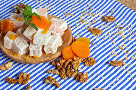 Close-up of turkish delight, lokum or rahat lokum, bright dried apricots on a wooden plate, leaves of mint, walnuts, crumbs of walnuts on the table on a striped background.の写真素材
