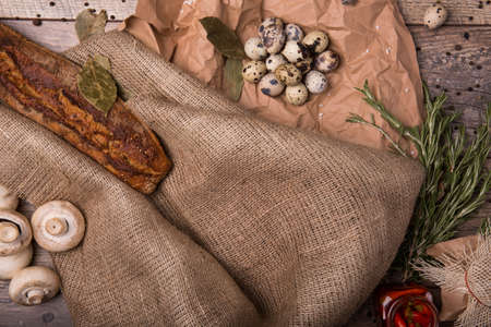 Top view of quail eggs on a brown paper and crusty, baked baguette on a burlap sack on a wooden background. White mushrooms and spices red hot chili pepper, black pepper and rosemary leaves.の写真素材