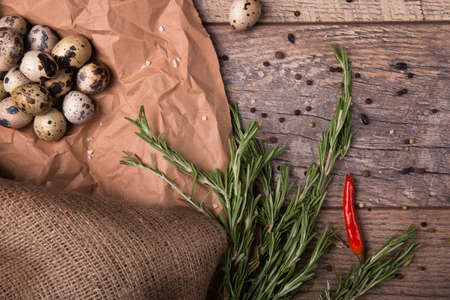 A view from above on a fresh, long rosemary leaves and bright red, spicy chili pepper on a brown wooden table background. Spices next to quail eggs on a crumpled paper. Healthy dinner preparation.の写真素材