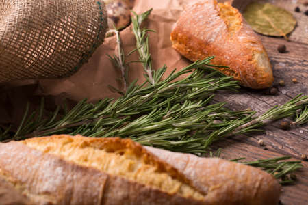 A close-up picture of a traditional french baguette on a dark brown wooden background. A beautiful fresh bread with long rosemary and dry bay leaves. Homemade spicy breakfast concept.の写真素材