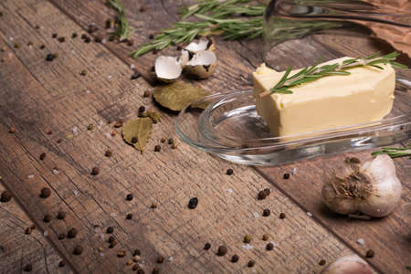 A close-up picture of butter in a glass container on a wooden table background. Egg shell and garlic. Rosemary twigs with black pepper peas and sea salt. Seasoning. Cooking concept.の写真素材
