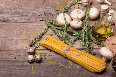 A view from above on a vegetable composition. Olive oil, garlic, rosemary twigs, pasta, quail eggs, and mushrooms. Raw ingredients on a wooden table background. A set of vegetables for dinner dishes.の写真素材