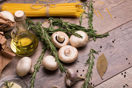 A beautiful uncooked dinner composition. A top view of Italian pasta, fragrant rosemary twigs, garlic, mushrooms and oil bottle. A set of raw ingredients for pasta dish on a wooden table background.の写真素材