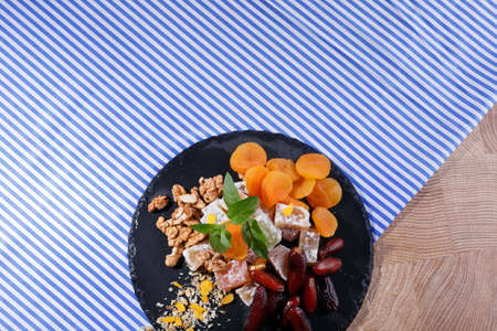 A close-up picture of a black plate with different confectionery ingredients on a striped fabric background. Orange apricots, nutritious walnuts, mint leaves, date fruits, turkish delight. Copy space.の写真素材