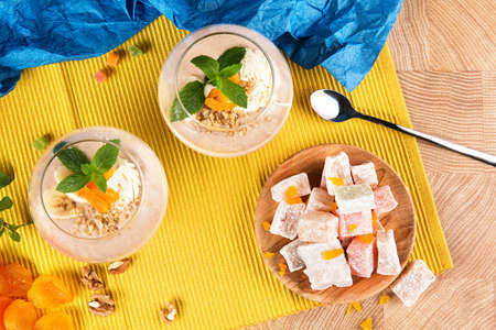 Two fruity smoothies in dessert glasses with sweets and fruits on a yellow background. Banana cocktails, Turkish Delight and dried apricots with walnuts on a colored background, top view.の写真素材