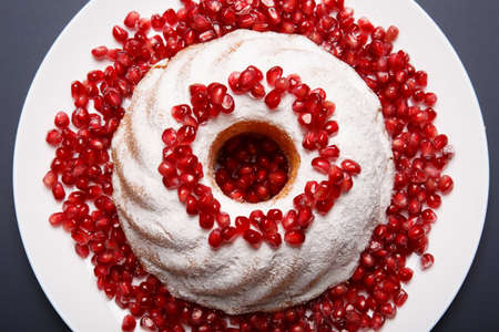 Top view of a perfectly round cake with sugary icing and decorative garnet seeds on a dark blue background. An appetizing, powdered ring cake on a white plate. Classic baked snacks for parties.の写真素材