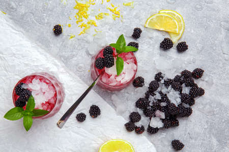 A couple of fancy glasses full of cold, icy beverage with spicy mint, lemon pieces, juicy blackberries and metal spoon on a frozen white table background. Refreshing cocktails with fruits and berries.の写真素材