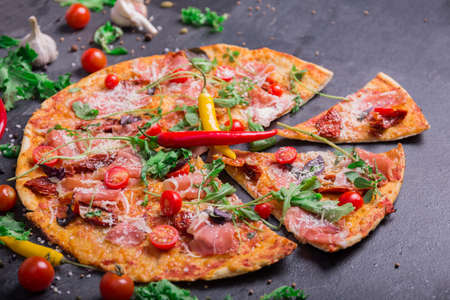 Close-up of a tasty pepperoni pizza with red hot chili pepper, meat, and spices on a dark stone background. Salad leaves, cherry tomatoes, garlic and peppercorns near a yummy meaty pizza.の写真素材