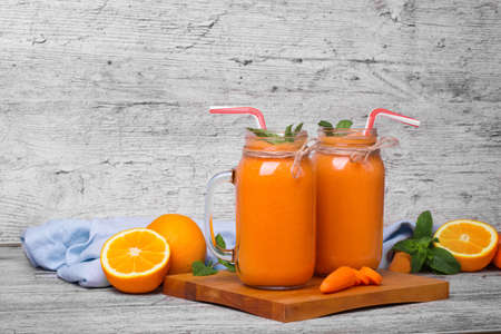 A pair of transparent jars full of nutritious, vegan orange vegetable juice with mint standing on a cutting desk. Chopped orange fruits and peeled carrots on a gray rustic background. Copy space.の写真素材