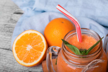 Close-up picture of a big bottle filled with orange carrot beverage with a striped straw on a decorative blue fabric background. Tropical oranges and aromatic mint next to a smoothie on a wooden desk.の写真素材