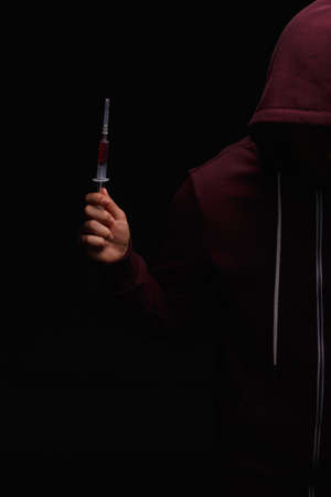 Photo of desperate young drug addict wearing a hood with a syringe in a hand on a black background. Drugs concept.の写真素材