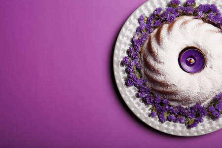 Top view of a ring cake covered with powdered sugar on a plate decorated with little purple flowers and an alight candle on a bright violet background, copy space.の写真素材