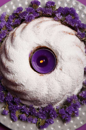 Close-up of a plate with a round cake, sprinkled with powdered sugar and a lit purple candle in the...