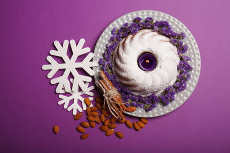 Top view of ring cake with an alight candle, cinnamon, almond and white snowflakes on a bright violet background.の写真素材