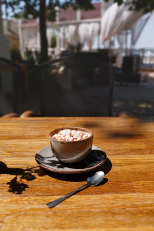 A close-up of a gray porcelain mug of sugar cappuccino with marshmallows. Sweet coffee drink on wooden table background. A cup with a round plate and a metal spoon. Copy space. Relaxation concept.の写真素材