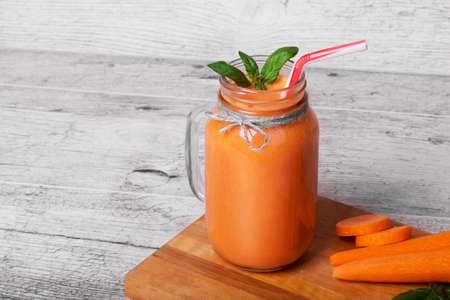 A refreshing drink next to raw peeled carrot tubes on a cutting desk. A mason jar full of detox beverage. A cool carrot smoothie with decorative basil leaves on a gray wooden background.の写真素材