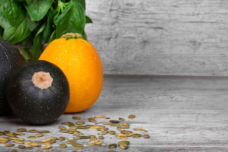 A wooden table with a green plant, vivid yellow and dark green zucchinis, seeds of squash, vegetables for vegetarian diet on a light wooden gray blurred background.の写真素材