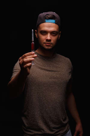A guy with a drug addiction holds a syringe with an injectable drug on a dark black background.