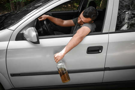 A drunk guy driving a car. A driving man with a bottle of alcoholic drink on an auto background. Alcoholism concept.の写真素材