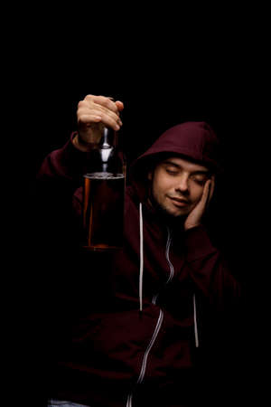 A drunk guy with a bottle of beer. Young alcoholic in a red hoodie on a black background. Alcoholic addiction concept.の写真素材