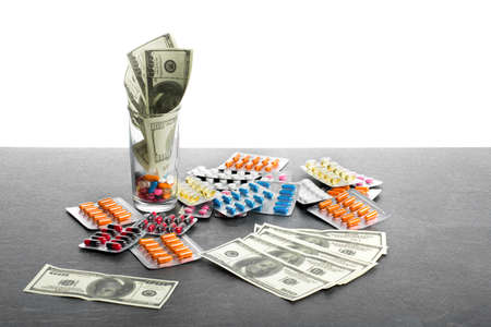 A close-up picture of a heap of colorful tablets, capsules, pills next to cash isolated on a white background. Hundred-dollar bills and drugs on a gray surface. Pharmacy, finance concept. Copy space.の写真素材