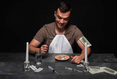 A smiling junkie with cash, drugs, and candles sitting at the table.A drug addict on a black background.  Copy space.の写真素材