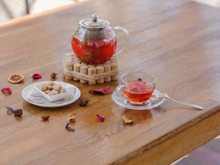 A bright berry tea in a glass teapot next to a transparent glass, a round white plate and dried fruits on a wooden background.の写真素材