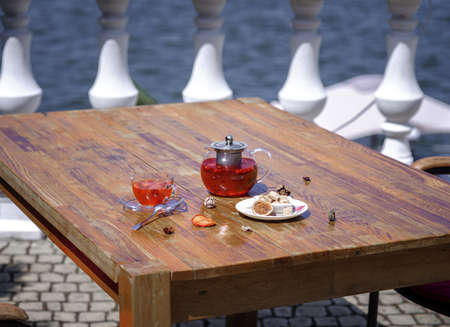 A beautiful bright composition of a teapot full of organic berry beverage on a brown wooden table. A plate with brown cubes of sugar, kettle and a cup of fresh wild strawberry tea on a sea background.の写真素材