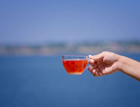 A ladys hand is holding a hot red drink from berries on a river blurred background. Fresh wild strawberry tea.の写真素材