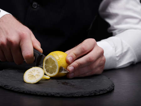 Close-up picture of bartender's hands cutting a ripe, raw, fresh yellow lemon on a black cutting desk on a bar counter background. Exotic fruity beverage preparation composition.の写真素材