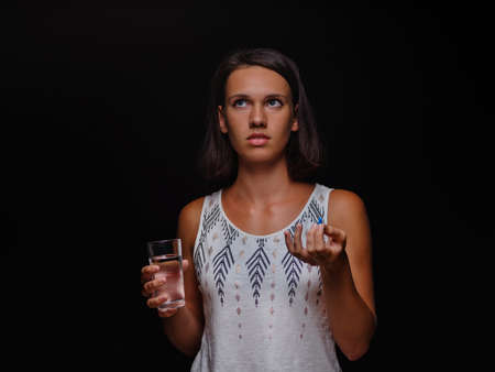 Young woman holding a glass of water and taking pills on a black background. Medical help, medicine concept. Copy space.の写真素材